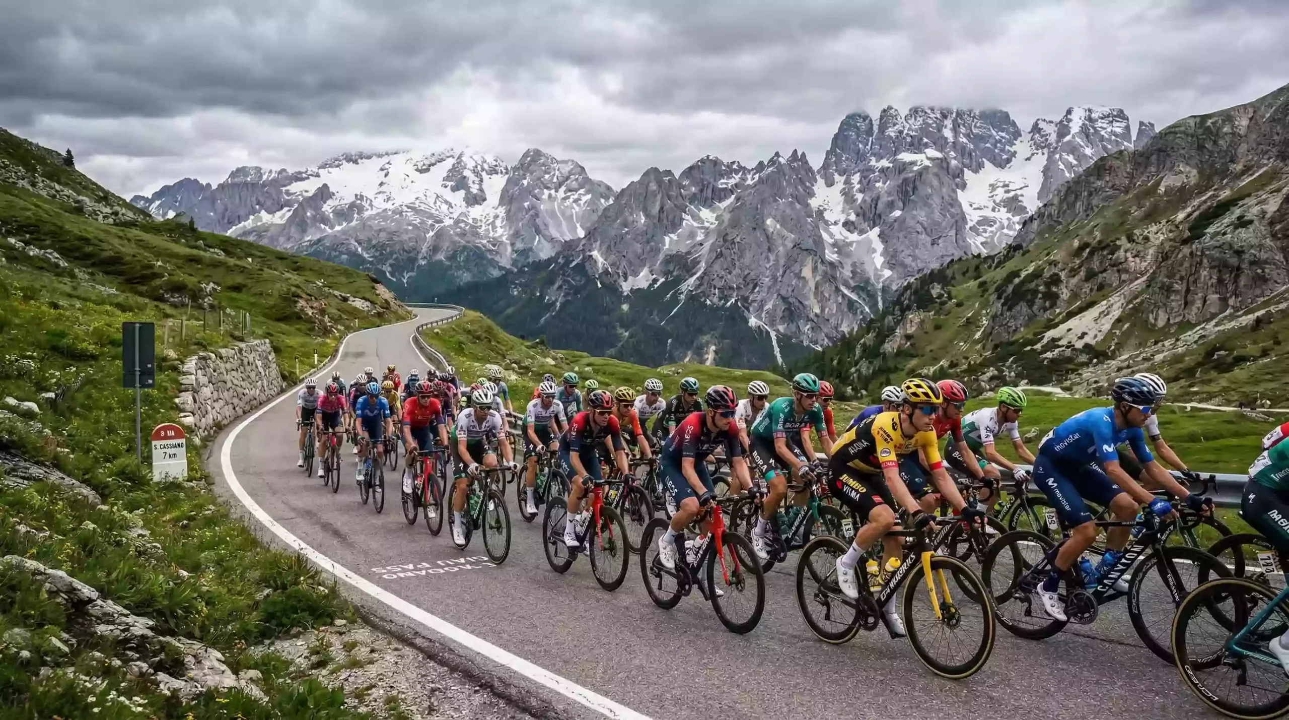 Pelotón ciclista ascendiendo un puerto de los Dolomitas italianos con nieve en las cumbres